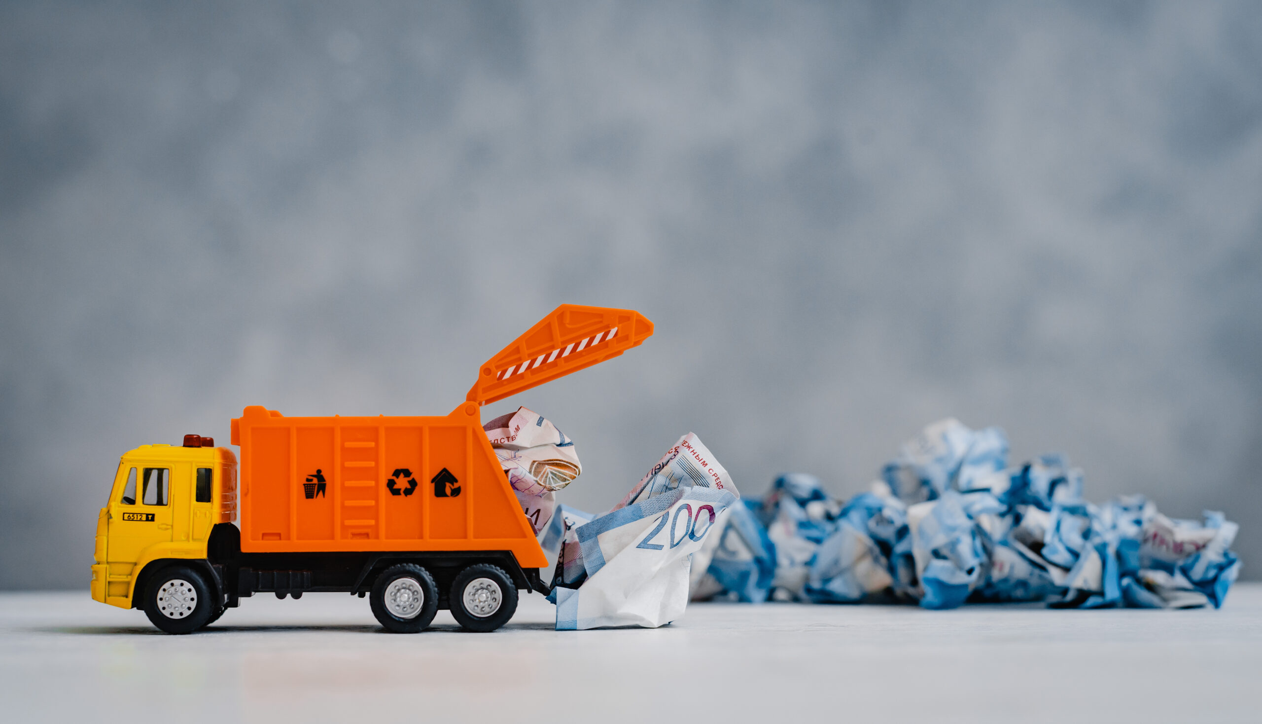 orange toy garbage truck unloads crumpled paper banknotes on a white table on a blue background.